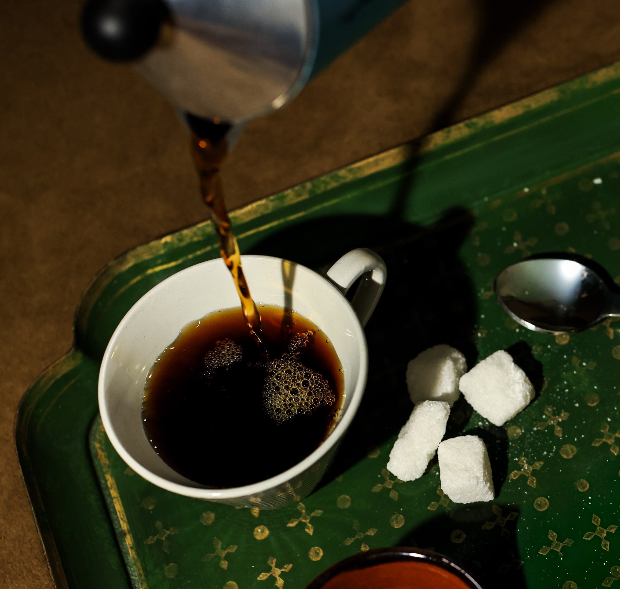 Coffee being poured into a cup on a green surface with sugar cubes and a spoon.